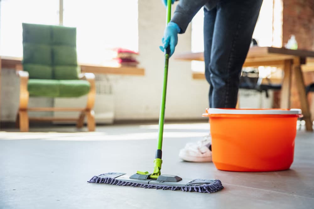 Person using a green mop to clean a floor next to an orange bucket in a bright room.